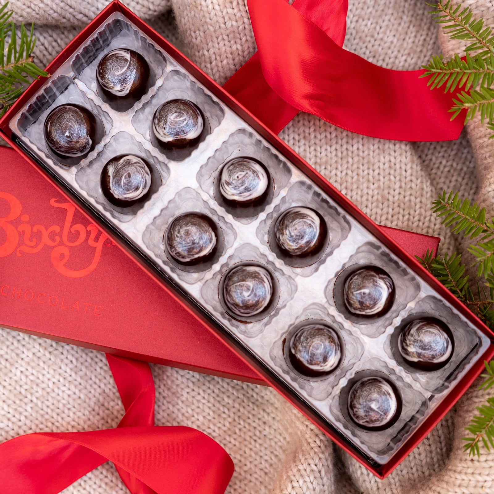 A red box of twelve round chocolates sits on a knitted beige blanket with red ribbons and green fir branches around it.