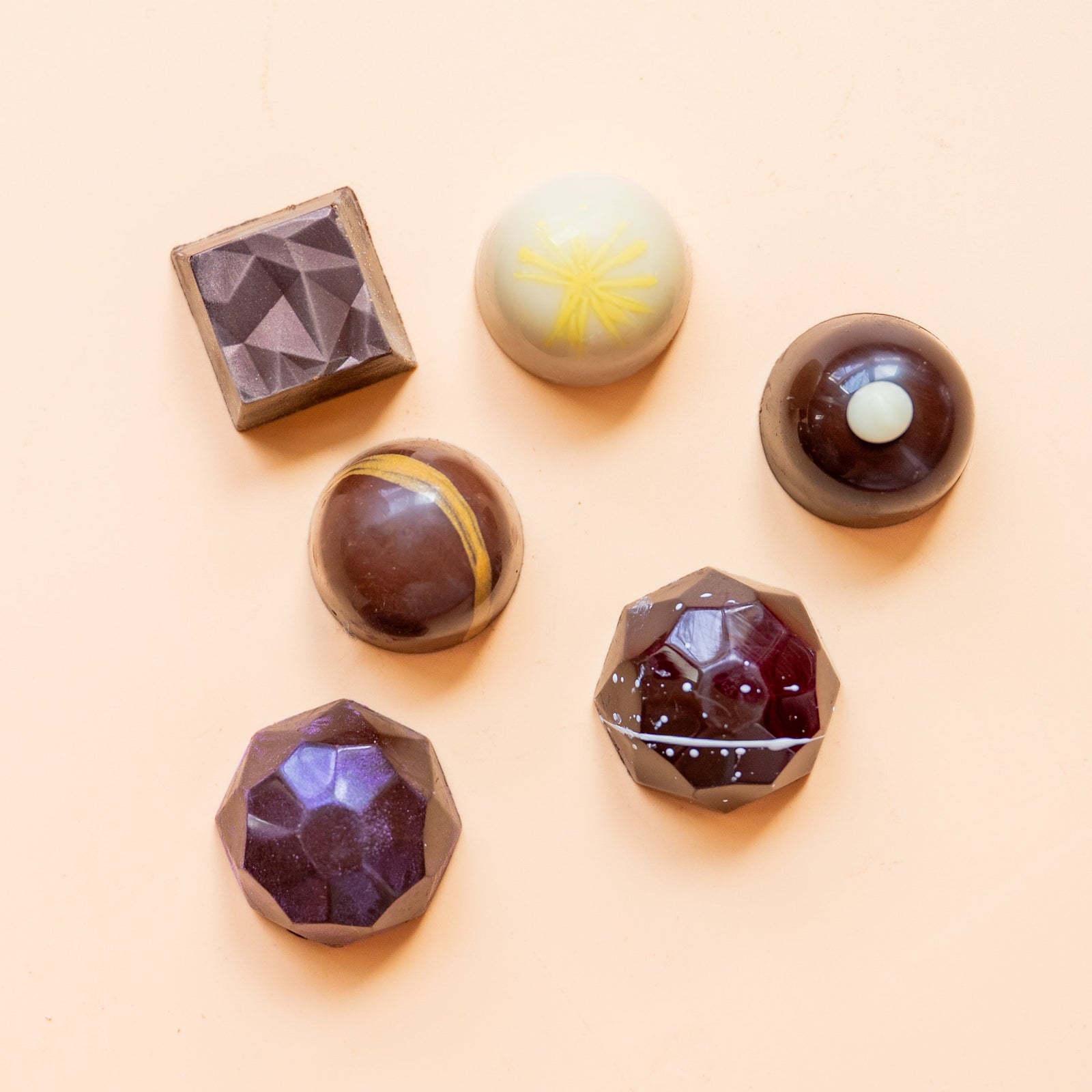 Six assorted artisan chocolates in geometric shapes and various glossy colors, arranged on a pale peach background.