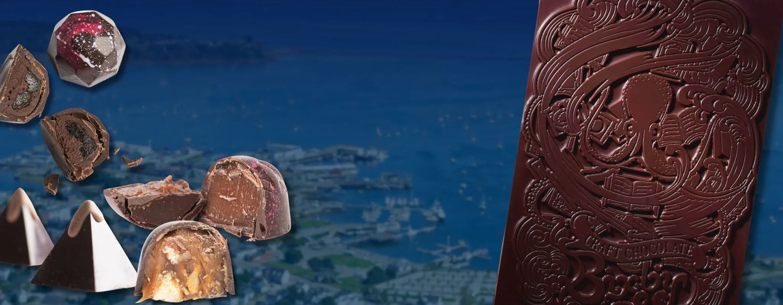 A variety of gourmet chocolates and truffles are displayed, with a large, intricately patterned chocolate bar on the right; a blurred cityscape and harbor can be seen in the background.