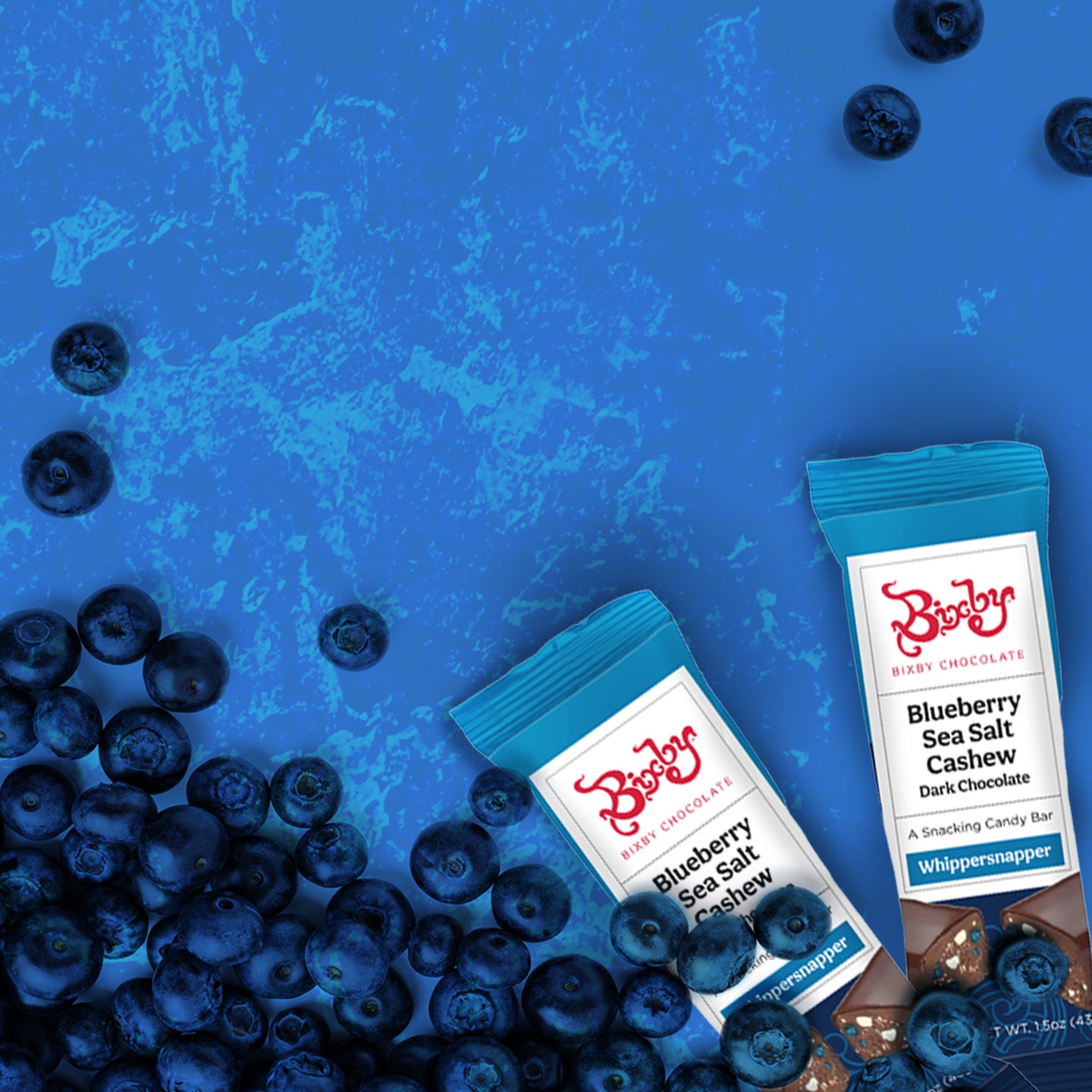 Two Bixby Chocolate Blueberry Sea Salt Cashew Dark Chocolate bars are on a blue surface surrounded by scattered fresh blueberries.