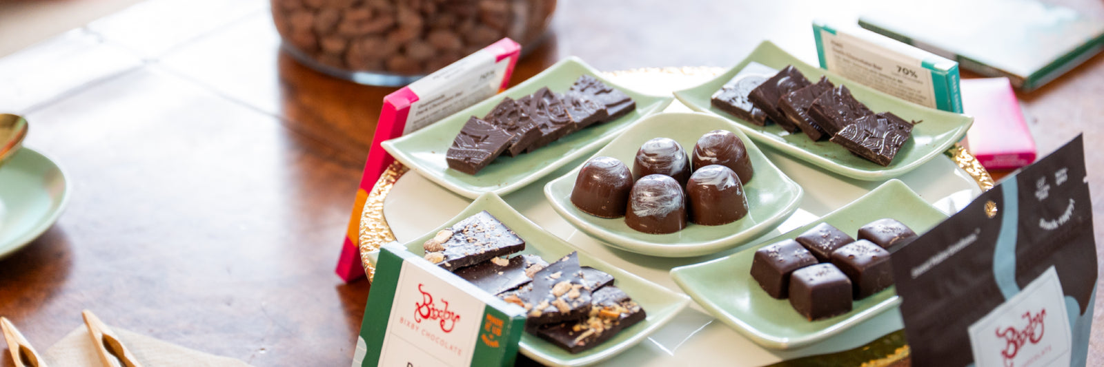 A selection of assorted dark chocolates and chocolate truffles is displayed on pale green plates, arranged on a round tray atop a wooden table, with packaged chocolate bars in the background.