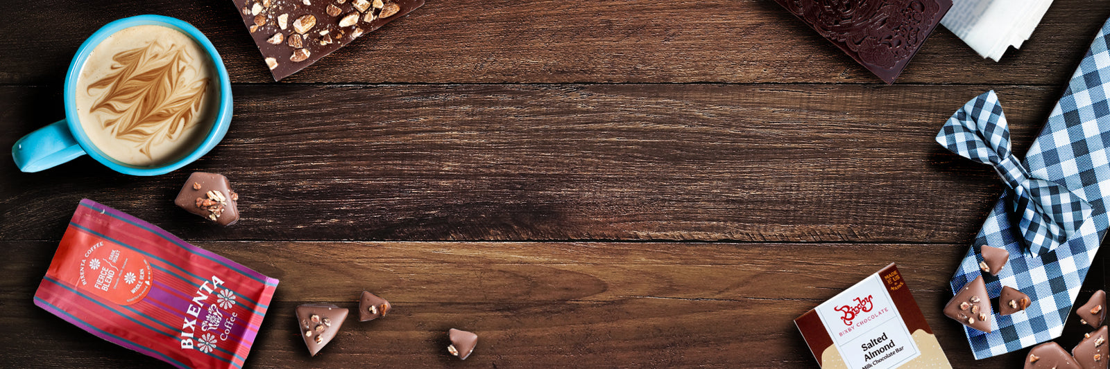 A blue cup of coffee, chocolate bars, small chocolate pieces, and a checkered cloth are arranged on a wooden table, leaving the center empty.