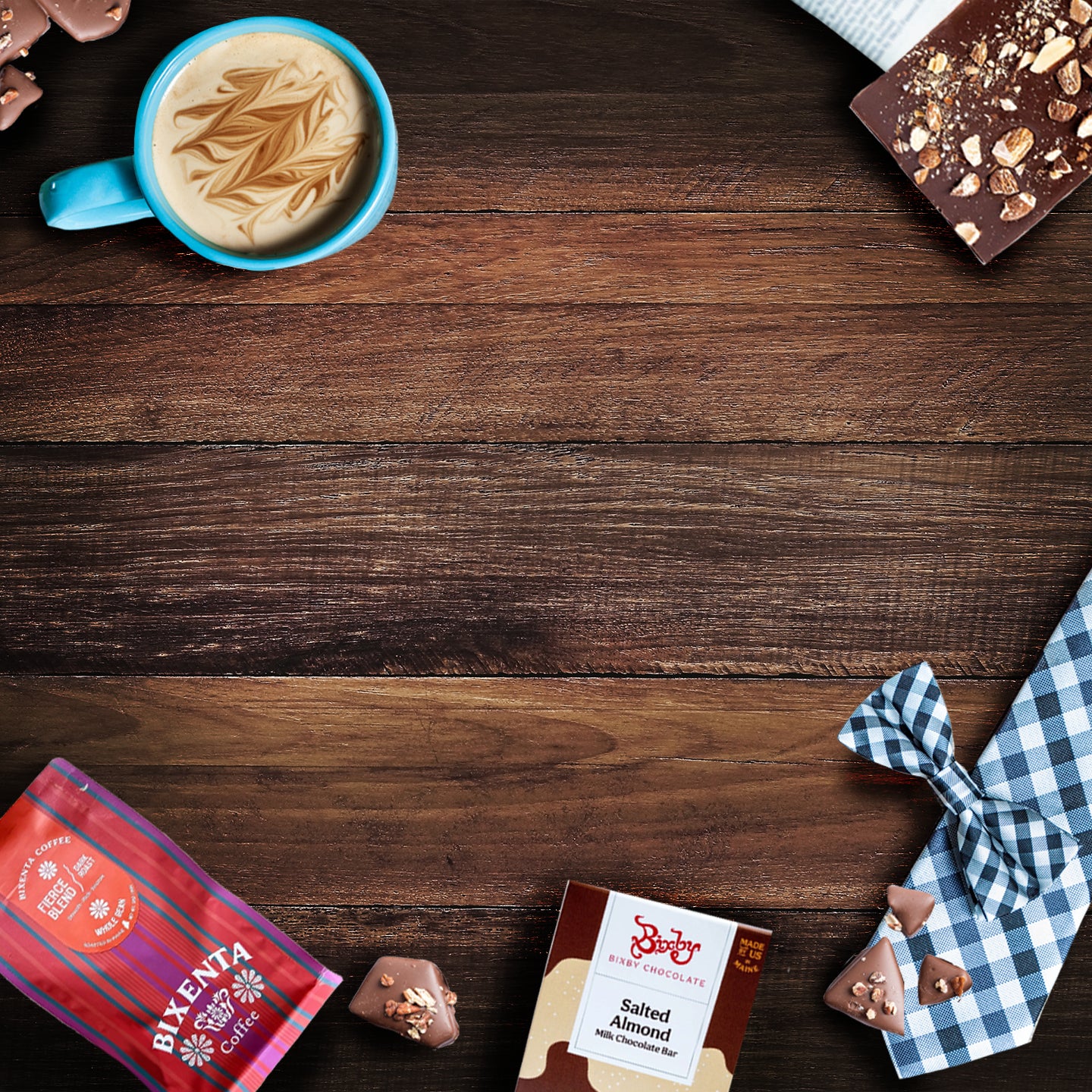A blue cup of coffee, a chocolate bar, chocolates, a red snack pack, a box of salted almond dark chocolate, and a blue gingham bowtie are arranged on a wooden surface.