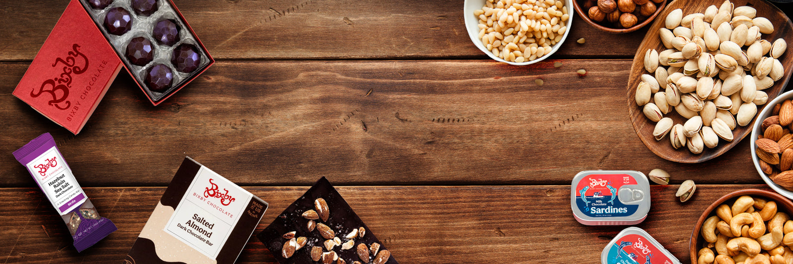 Assorted nuts and chocolates in bowls and boxes, along with two tins of sardines, are arranged on a wooden table with empty space in the center.