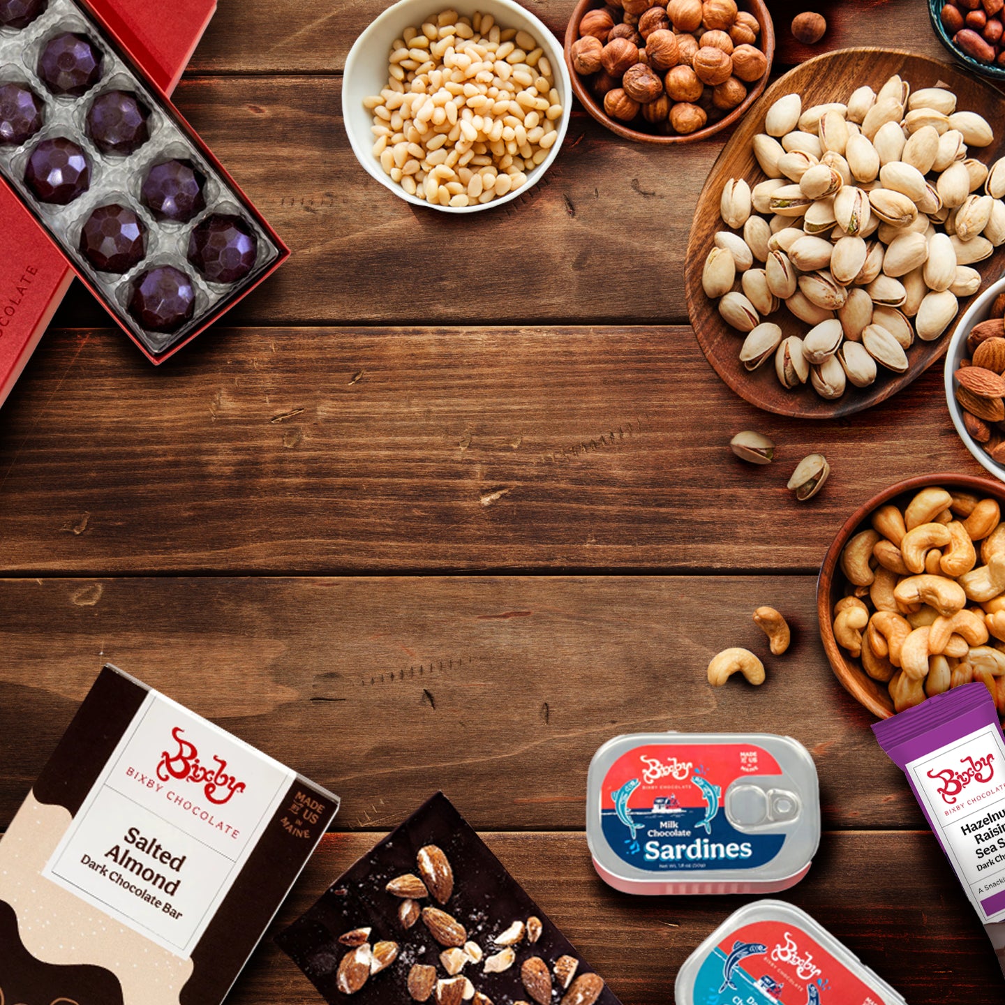 A wooden table with various snacks, including chocolates, bowls of pistachios, pine nuts, hazelnuts, and cashews, tins of sardines, and a package of salted almond dark chocolate bars.