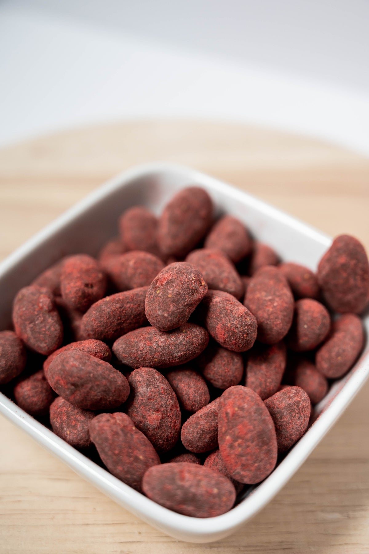 A white square bowl of Bixby Chocolates Strawberries & Cream Almonds sits on a light wooden surface, their red, powder-coated shells evoking the flavor of strawberry milk chocolate.