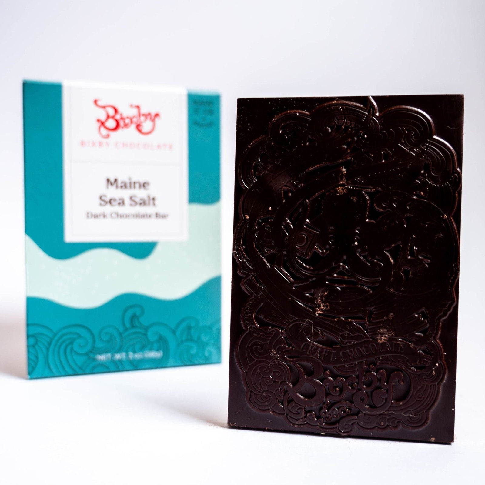 A dark chocolate bar with an ornate design stands upright before turquoise and white packaging labeled “Bixby Chocolates Organic Dark Chocolate Maine Sea Salt Bar,” highlighting organic chocolate and Maine sea salt flavors.
