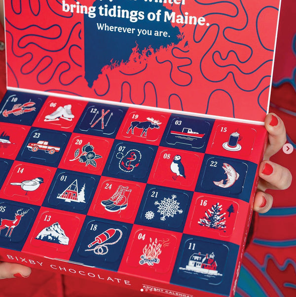 A person with red-painted nails holds an open Bixby Chocolate advent calendar decorated with winter and Maine-themed illustrations, such as a moose, sled, lighthouse, lobster, pine tree, and snowflakes.
