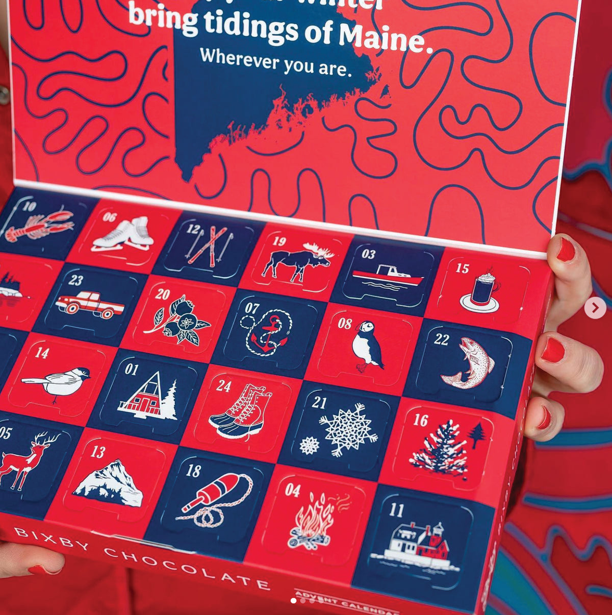 A person with red-painted nails holds an open Bixby Chocolate advent calendar decorated with winter and Maine-themed illustrations, such as a moose, sled, lighthouse, lobster, pine tree, and snowflakes.