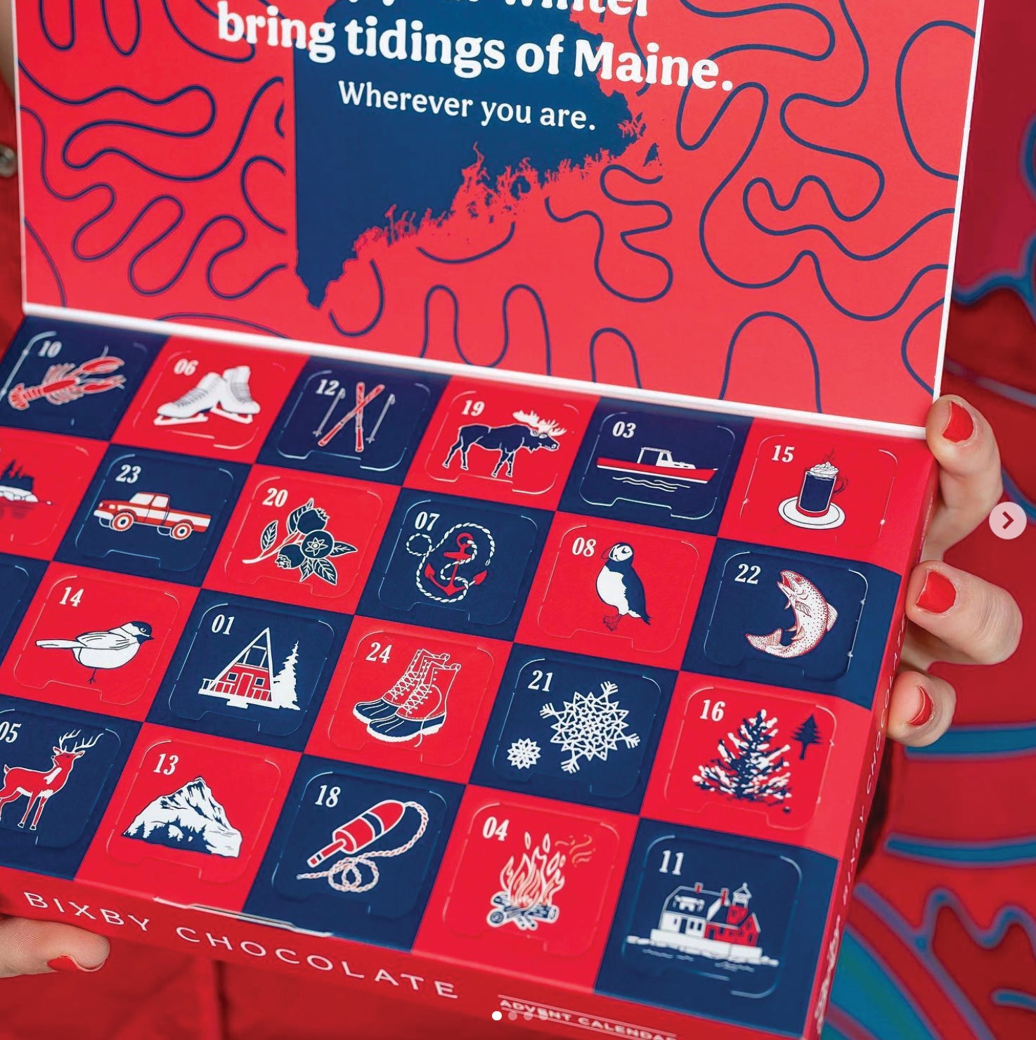A person with red-painted nails holds an open Bixby Chocolate advent calendar decorated with winter and Maine-themed illustrations, such as a moose, sled, lighthouse, lobster, pine tree, and snowflakes.