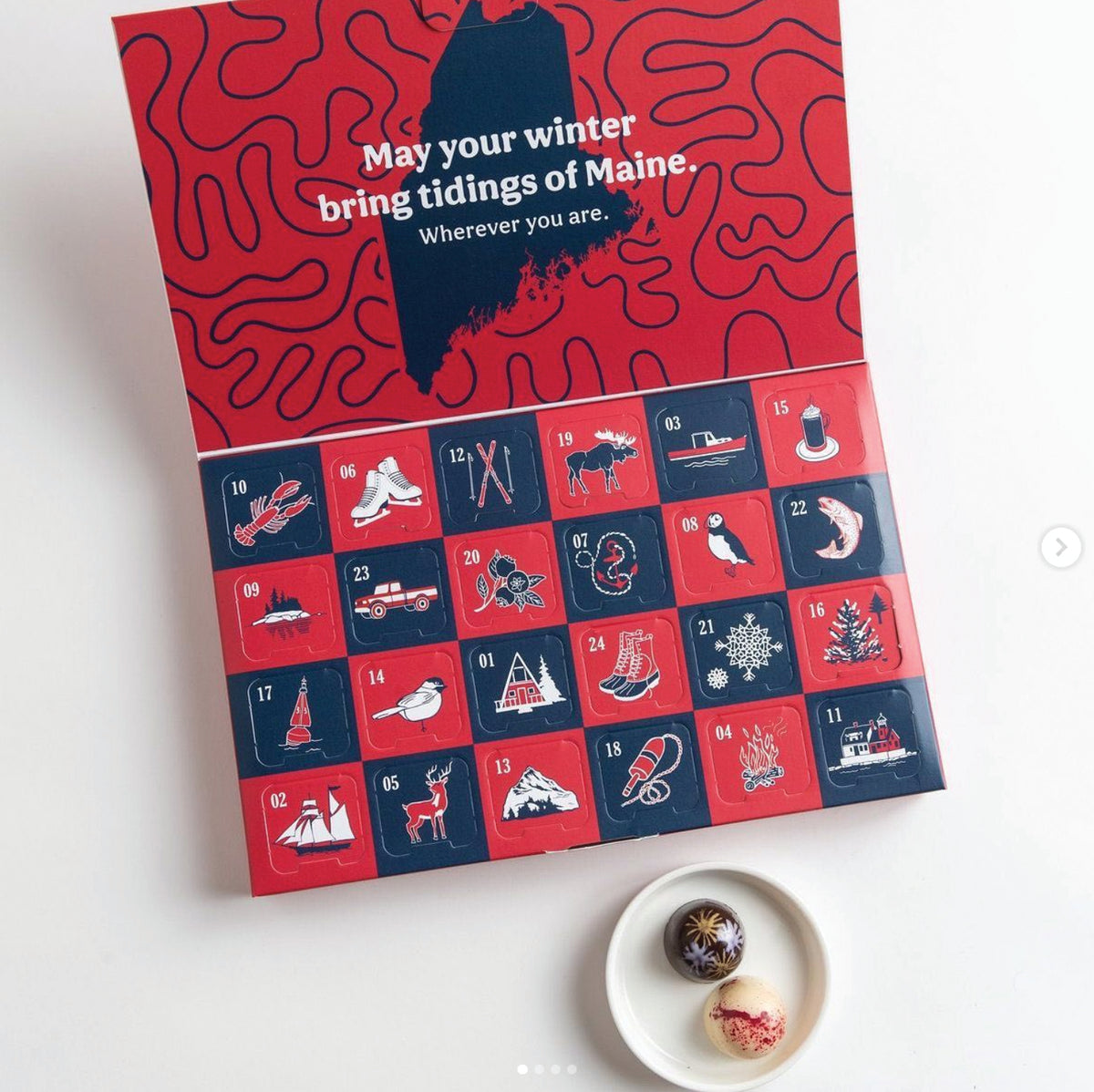 A red and navy advent calendar with Maine-themed illustrations and the text, “May your winter bring tidings of Maine. Wherever you are.” Two decorated chocolate truffles sit on a small white plate below the calendar.