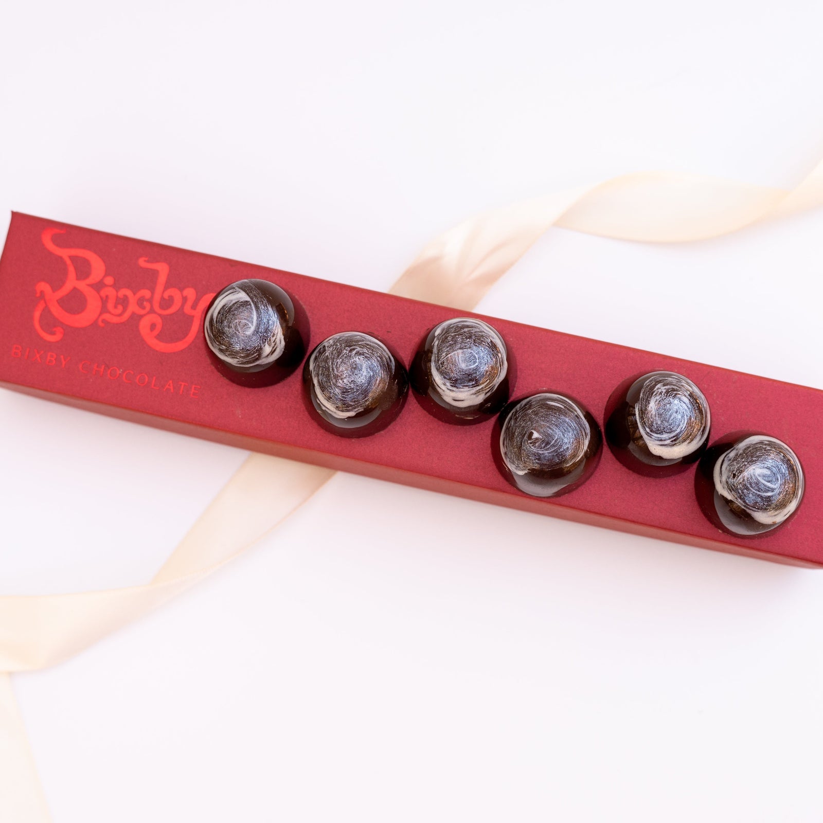 A red box labeled Bixby Chocolates holds six glossy Eggnog Bon Bons with a marbled silver and dark design. A cream ribbon is loosely arranged around the box on a white surface.