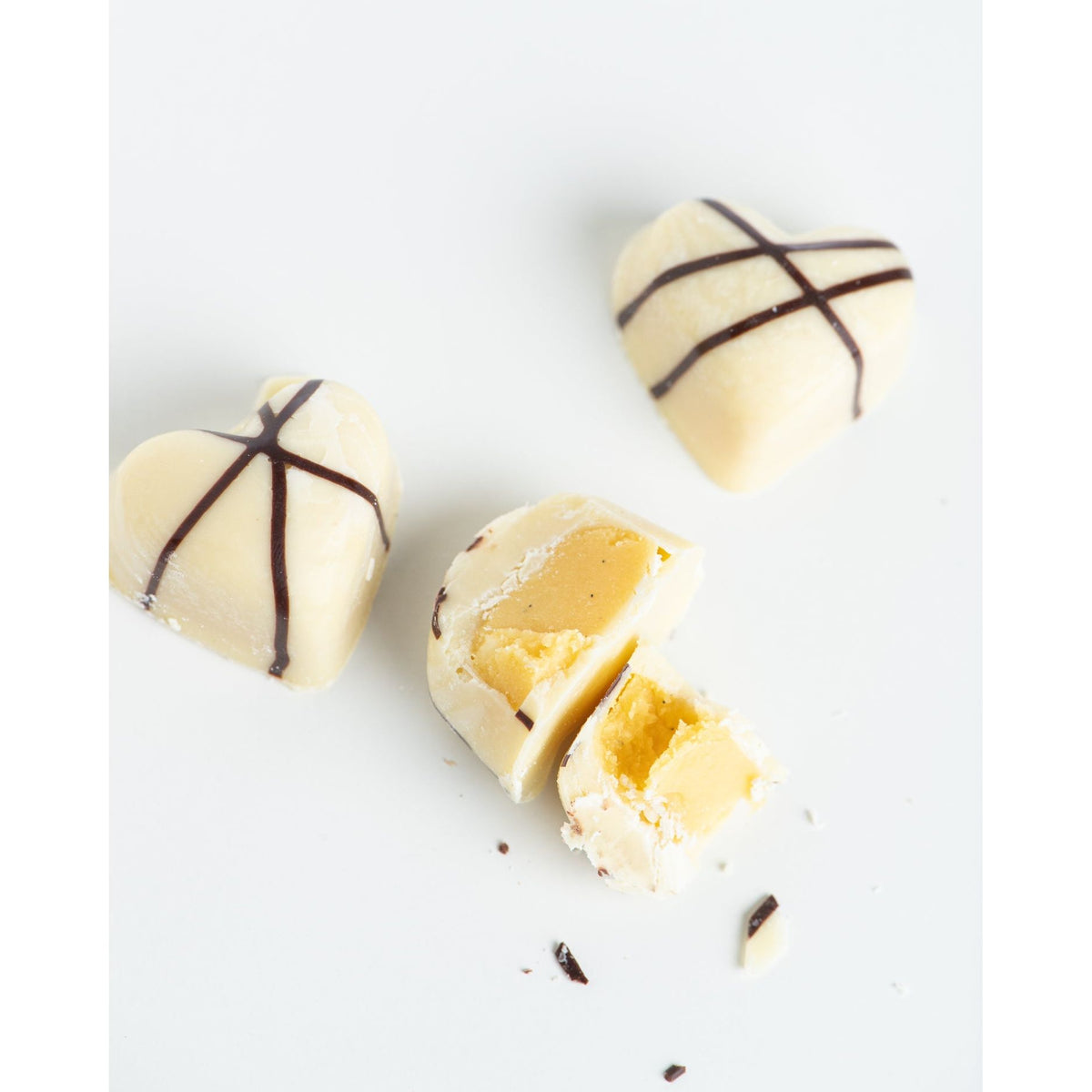 “3 Months of Chocolate Heart Love” by Bixby Chocolates features three heart-shaped bon bons with dark chocolate stripes, one opened to reveal creamy passion fruit ganache, all on a white surface with chocolate fragments.