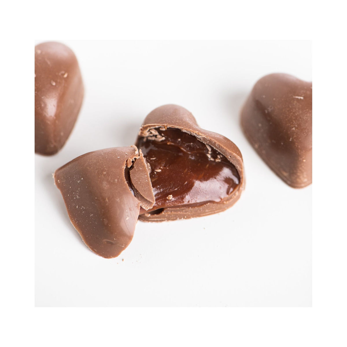 Three heart-shaped bon bons from Bixby Chocolates’ 3 Months of Chocolate Heart Love rest on a white surface, one broken open to reveal a glossy passion fruit ganache filling inside.