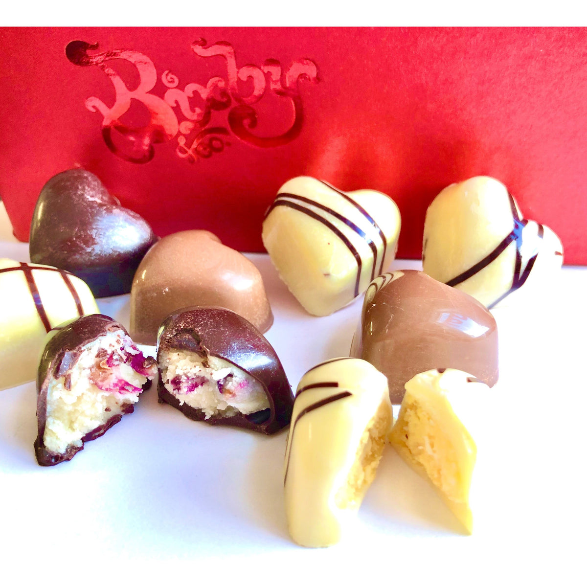 An array of heart-shaped chocolates—white, milk, and dark with unique fillings—is displayed in front of a red Bixby Chocolates box, representing the "3 Months of Chocolate Heart Love" gift set.
