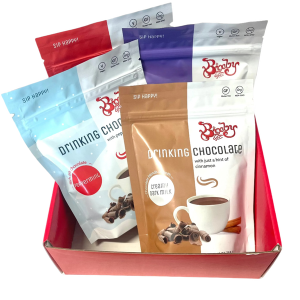 The Bixby Chocolates Drinking Chocolate Gift Box features four colorful packets of chocolate mixes in flavors like peppermint and creamy dark milk, each bag illustrated with chocolate ingredients—a perfect red gift box.