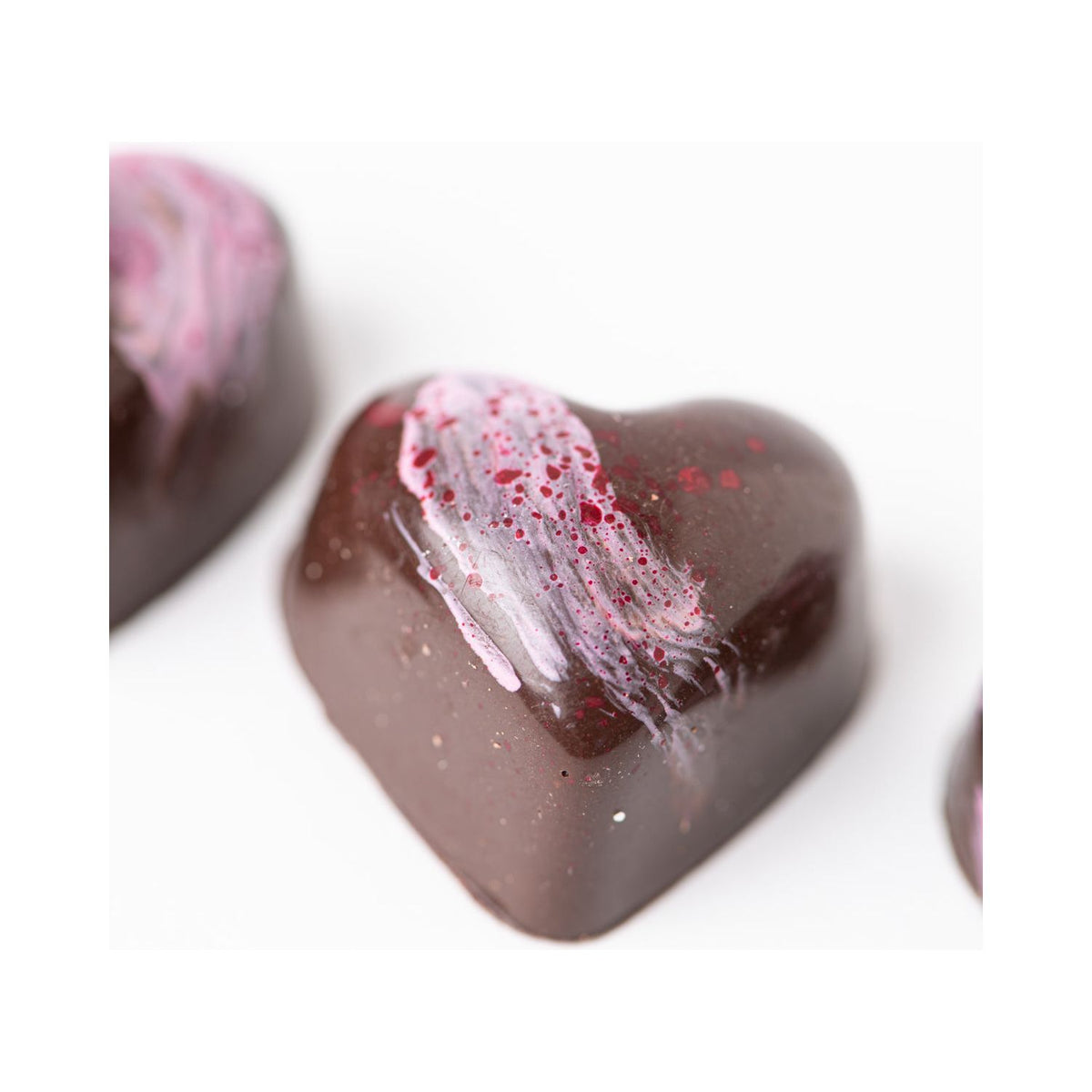 The Bixby Chocolates "3 Months of Chocolate Heart Love" features glossy, heart-shaped dark chocolates with pink and red splashes. Two at the edges reveal hints of blood orange caramel or passion fruit ganache on a white background.