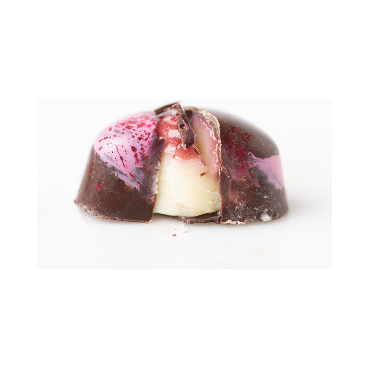 A close-up of Bixby Chocolates’ 3 Months of Chocolate Heart Love: a glossy dark bonbon sliced open to reveal passion fruit ganache and blood orange caramel, with pink and red splashes on a white background.