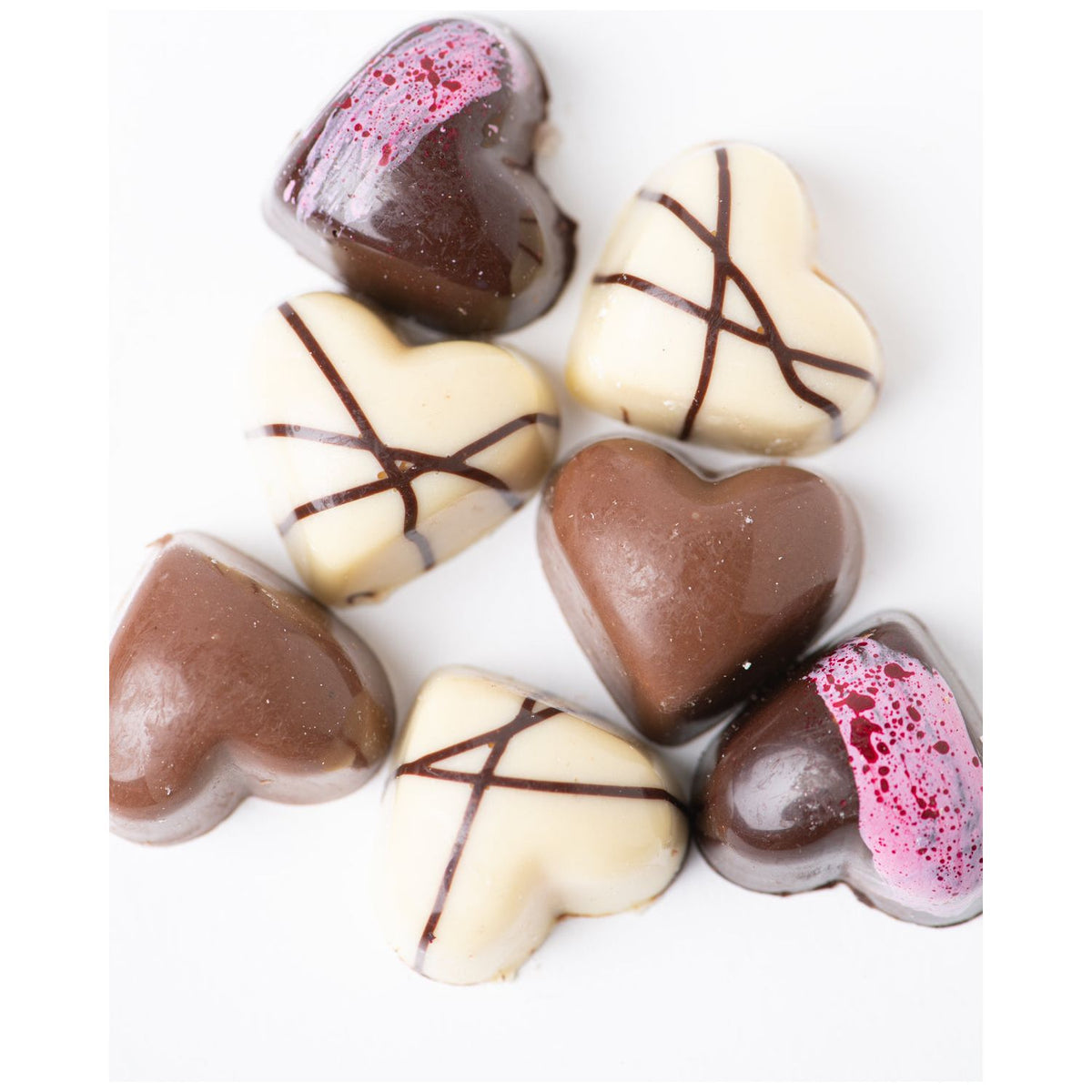 Bixby Chocolates’ 3 Months of Chocolate Heart Love showcases seven heart-shaped bon bons in white, milk, and dark chocolate—some with passion fruit ganache—arranged in a loose cluster on a white background.