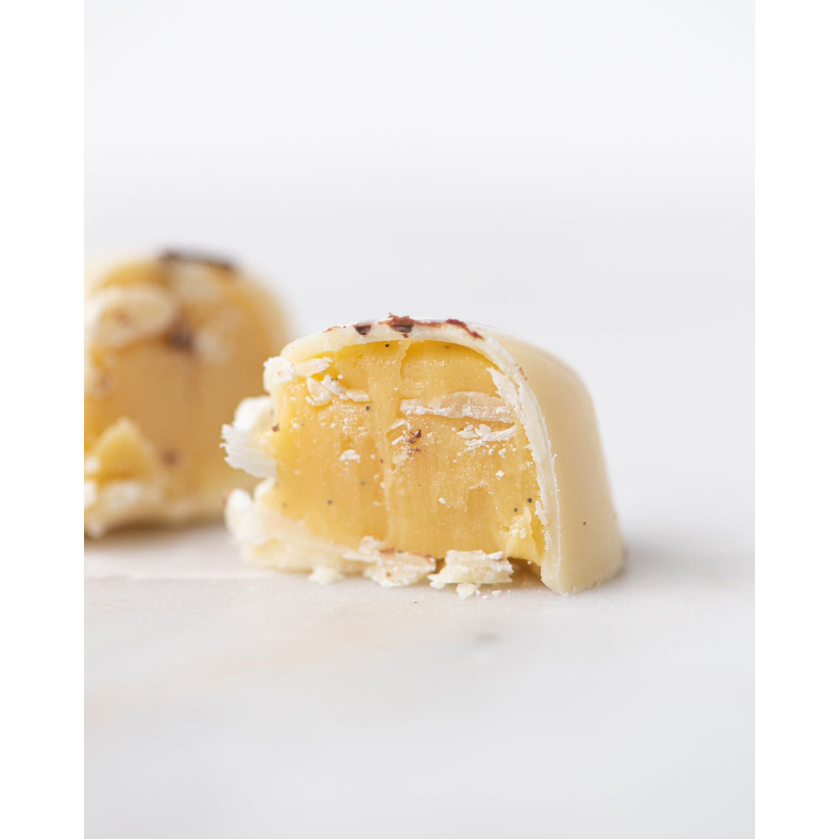 A white chocolate truffle from Bixby Chocolates' 3 Months of Chocolate Heart Love, broken in half to reveal its creamy passion fruit ganache, sits on a white surface with another half-blurred truffle behind it.