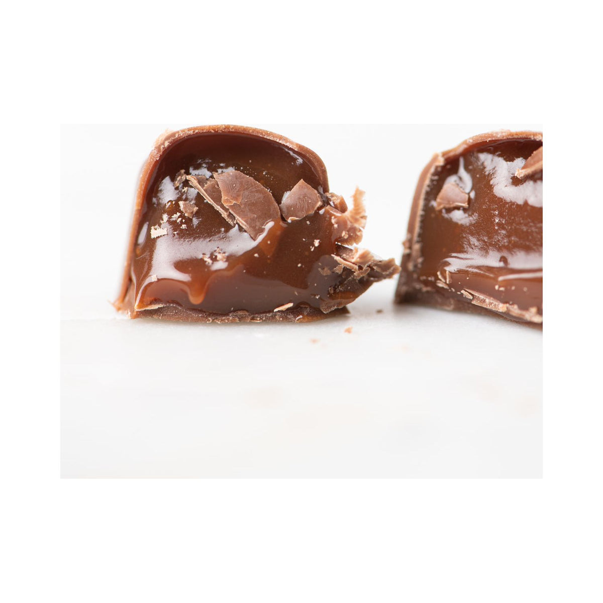 Close-up of Bixby Chocolates’ 3 Months of Chocolate Heart Love: a chocolate candy cut in half reveals smooth, gooey blood orange caramel filling with bits of milk chocolate and caramel oozing out on a white background.