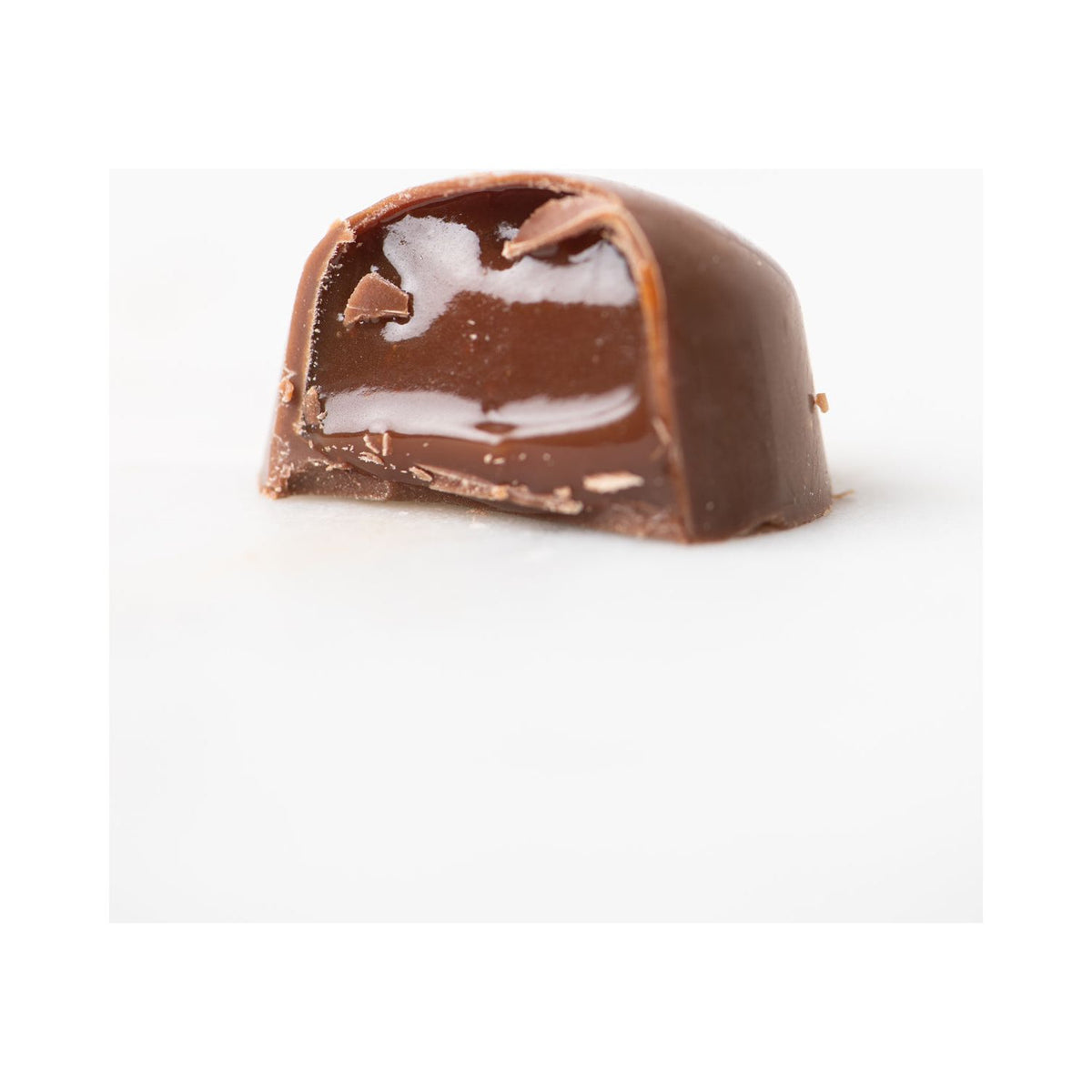 A close-up of Bixby Chocolates' 3 Months of Chocolate Heart Love shows a heart-shaped bon bon cut in half, revealing creamy white and brown layered filling, set against a plain white background.